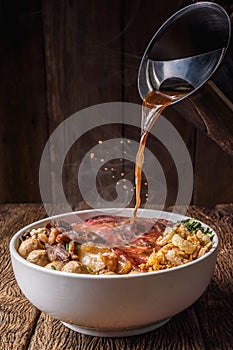 Bowl of Thai style beef noodle with assortment of beef and chinese thick soup pouring from soup jar