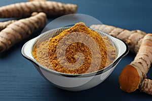 Bowl with turmeric powder and raw roots on blue wooden table, closeup