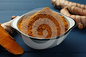 Bowl with turmeric powder and raw roots on blue wooden table, closeup