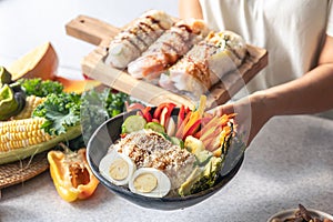 Bowl with vegetables and rice and spring rolls in female hands in the kitchen.