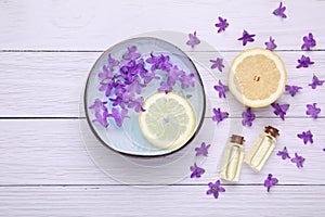 Bowl with water, lemon, purple flowers and bottles of essential oils on white wooden table, flat lay. Spa composition