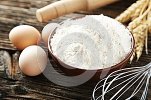 Bowl of wheat flour with eggs and whisk on brown wooden background