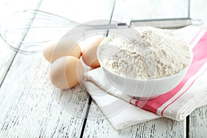 Bowl of wheat flour with eggs and whisk on white wooden background