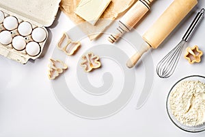 Bowl with wheat flour, rolling pin, whisk, eggs, butter, cookie cutters. Top view on a white table with a copy space