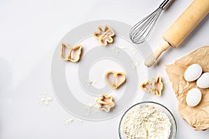 Bowl with wheat flour, rolling pin, whisk, eggs, cookie cutters. Top view on a white table with a copy space