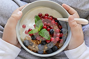 A bowl with yogurt, granola, blueberries and cranberries in the hands of a child, healthy breakfast concept.