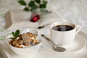 A bowl of yogurt and granola, a cup of coffee on a white tray on the bed, in the background an open book with a red rose