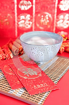 A bowl of Yuanxiao and hongbao in red background. The Chinese characters in the picture mean `happiness` and `good luck`