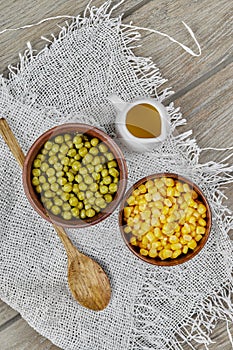 Bowls of boiled sweet corn and green peas, a sauce, and a wooden spoon on a wooden table