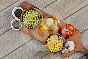 Bowls of boiled sweet corn and green peas, spices, oil, and vegetables on a wooden board