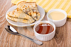 Bowls with condensed milk, apricot jam, napkin, folded pancakes on saucer, spoon on table