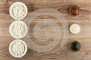 Bowls with cottage cheese, bowls with boiled condensed milk, condensed milk, gooseberry jam, and spoons on table. Top view