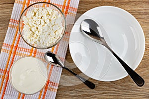 Bowls with cottage cheese, sour cream, spoon on napkin, spoon in plate on table. Top view
