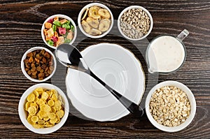 Bowls with dried fruits, corn flakes, oatmeal, sunflower seeds, banana chips, yogurt, bowl and spoon on table. Top view