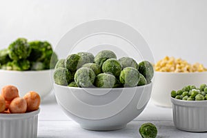 Bowls of frozen vegetables: brussels sprouts, peas, broccoli, corn, peas, carrots on a white plate.