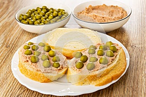 Bowls with green peas and liver pate, sandwiches with pate and green peas, slice of bread in plate on table