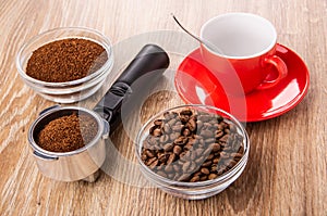 Bowls with ground coffee and coffee beans, holder from coffee maker with coffee, spoon in cup on saucer on table