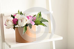 Box with beautiful peony flowers on shelf near white wall