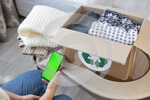 A box of clothes with a recycling sign sits on a table in the living room. A woman sits next to her and prints an