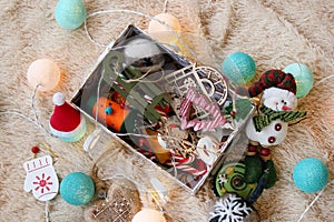 Box with colorful wooden and felt Christmas decorations, caramel sticks and Christmas lights on a soft beige blanket.