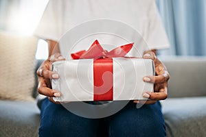 Box, gift and woman on a sofa in the living room with a giving gesture for celebration or event. Bow, wrapping paper and