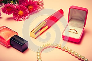 Box with golden ring, lipstick, nail polish and flowers on a pink background