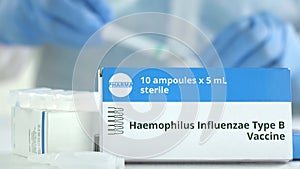 Box with haemophilus influenzae type B vaccine on the table against blurred lab assistant. Fictional phaceutical logo photo
