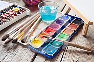 Box of watercolor paints, art brushes, glass of water and easel