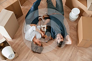 Between boxes, lying down. Young couple with dog are moving to new home