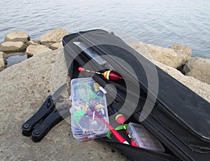 boxes made of translucent plastic with floats for multitool fishing tackle and a bag for a fishing rod lie on a large stone