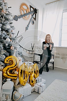 Boxing day. Christmas gift ideas for woman. One redhead girl in black evening dress unpack gifts near Christmas tree at home