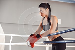 Boxing. Woman in gloves boxer leaning on rope on ring looking aside curious