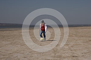 A boy of 10 years old in a white sweatshirt and orange vest plays football on a deserted beach in solitude