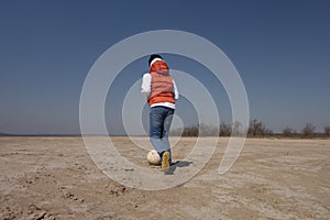 A boy of 10 years old in a white sweatshirt and orange vest plays football on a deserted beach in solitude