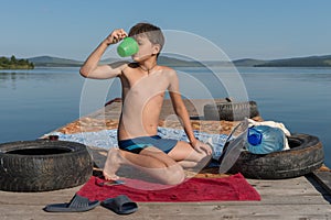 A boy of 11 years old slake his thirst with water from a mug, sitting on a wooden pier, against the backdrop of the lake on a