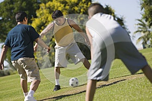 Boy (13-15) with two young men playing soccer outdoors in park.