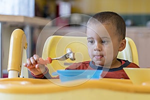 The boy 2 years eating meat. Children`s table. The concept of the child`s independence. funny kid in a baby seat.