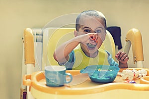 The boy 2 years eats porridge. Children`s table. The concept of the child`s independence. Cute toddler boy with blue spoon is