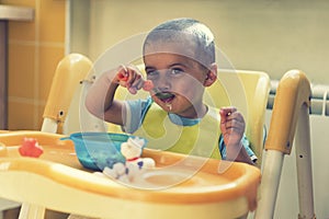 The boy 2 years eats porridge. Children`s table. The concept of the child`s independence. funny kid in a baby seat. toned