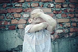 A boy of 4-5 years old covered his face with his hands and is sad against the background of a brick wall. Child and emotional expe