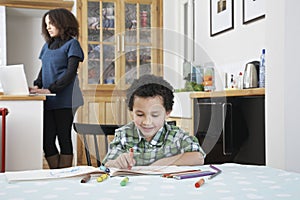 Boy (5-6) colouring in colouring book mother using laptop in background