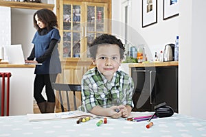 Boy (5-6) colouring in colouring book mother using laptop in background