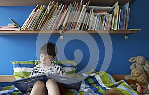 Boy (5-6) sitting on bed reading book