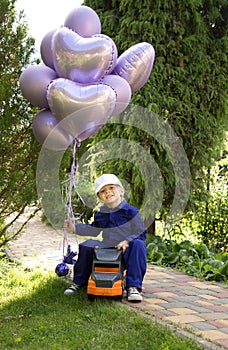 boy of 5-6 years old is sitting cheerfully on a large toy car, holding a bunch of purple balloons