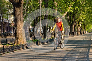A boy of 8 years old in an orange vest rides a bicycle in a city park on a sunny spring morning