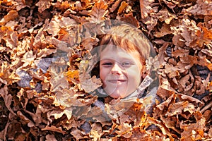 Boy 9-10 years old lies buried in autumn yellow oak leaves in park in rays of setting sun and smiles.