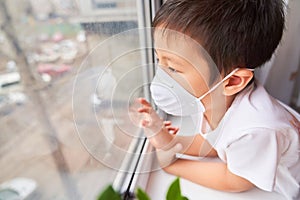 A boy of Asian appearance is standing in a white medical mask by the window, self-isolation - a child in a mask looks at the