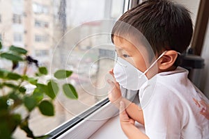 A boy of Asian appearance is standing in a white medical mask by the window, self-isolation - a child in a mask looks at the