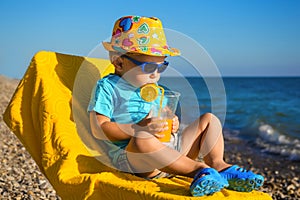 Boy baby in sun glasses and hat on beach drinks juice