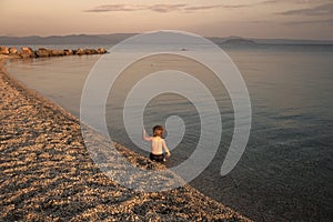 Boy on the beach. concept of loneliness. Cute child waving hand at seashore in evening. Boy sits in sea water, rear view